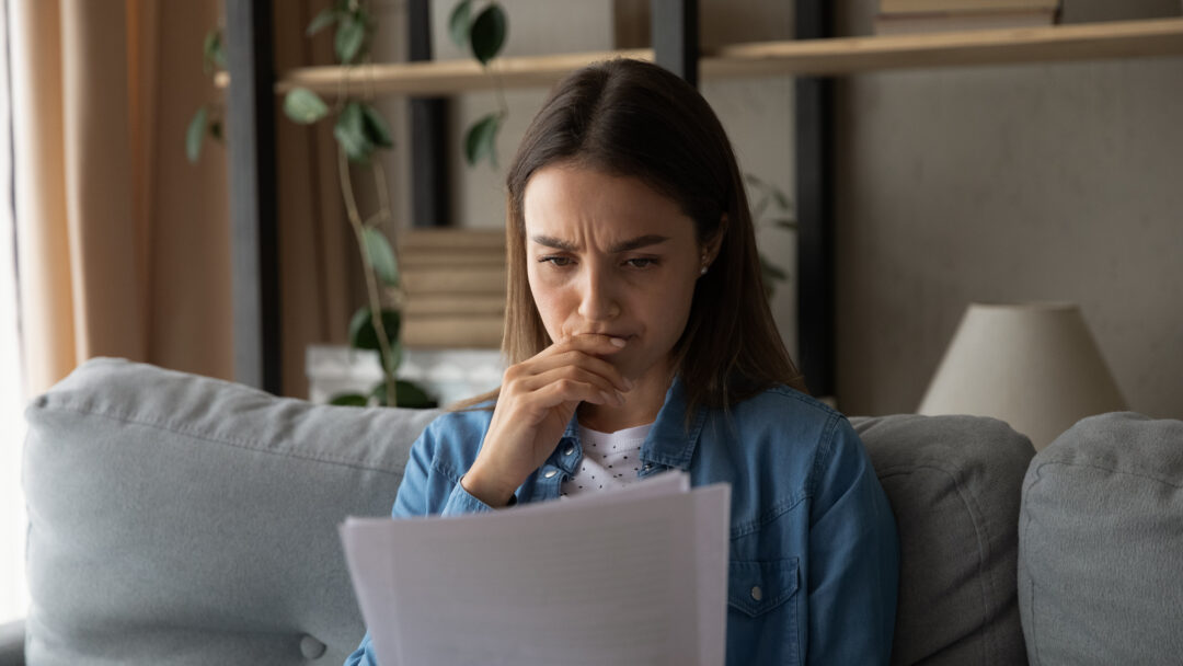 Person sitting on a couch reading a document with a concerned expression, hand near their mouth