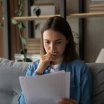 Person sitting on a couch reading a document with a concerned expression, hand near their mouth
