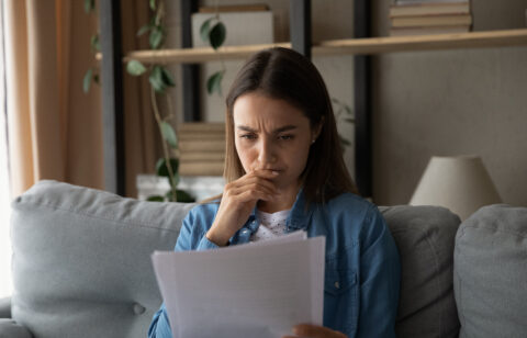 Person sitting on a couch reading a document with a concerned expression, hand near their mouth