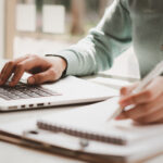 Close-up of hands typing on a laptop while writing notes in a notebook