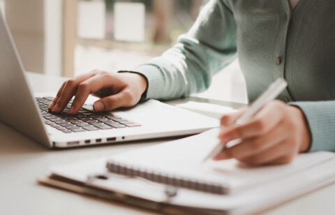 Close-up of hands typing on a laptop while writing notes in a notebook