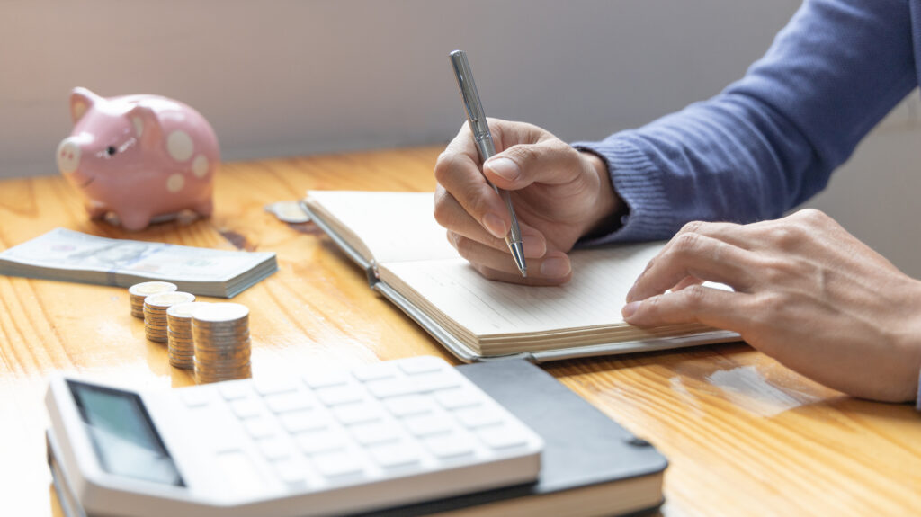 Person writing in a budgeting notebook beside a calculator, coins, cash, and a piggy bank on a wooden table.