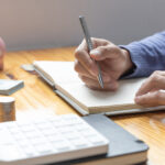 Person writing in a budgeting notebook beside a calculator, coins, cash, and a piggy bank on a wooden table.