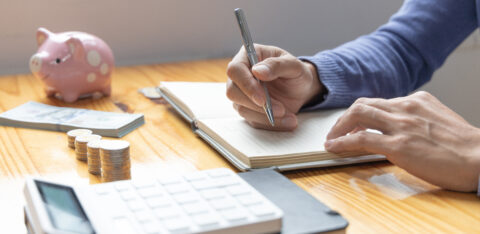 Person writing in a budgeting notebook beside a calculator, coins, cash, and a piggy bank on a wooden table.