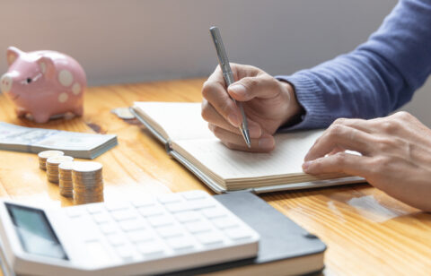 Person writing in a budgeting notebook beside a calculator, coins, cash, and a piggy bank on a wooden table.