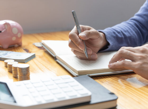 Person writing in a budgeting notebook beside a calculator, coins, cash, and a piggy bank on a wooden table.