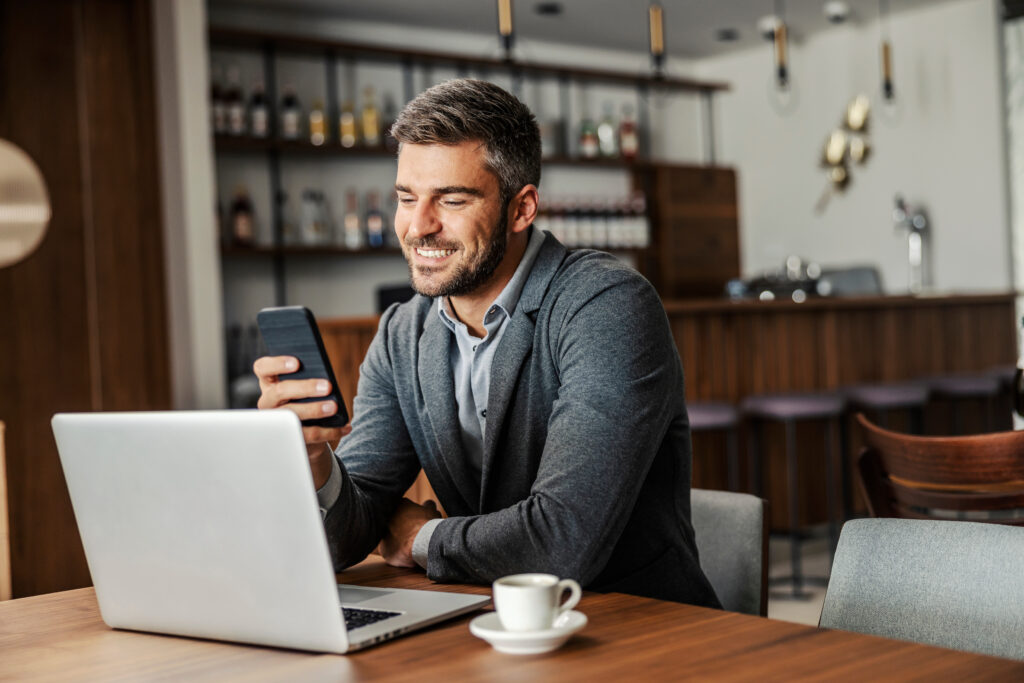 Person sitting at a table using a laptop while looking at a smartphone, with a coffee cup nearby