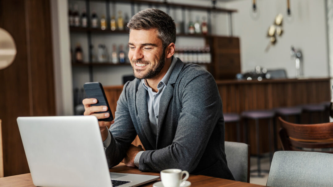 Person sitting at a table using a laptop while looking at a smartphone, with a coffee cup nearby