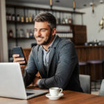 Person sitting at a table using a laptop while looking at a smartphone, with a coffee cup nearby