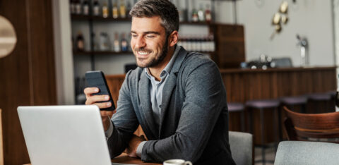 Person sitting at a table using a laptop while looking at a smartphone, with a coffee cup nearby