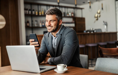 Person sitting at a table using a laptop while looking at a smartphone, with a coffee cup nearby