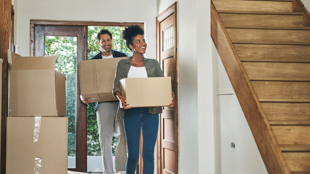 Couple carrying moving boxes into a home, stepping through the front door with more boxes nearby