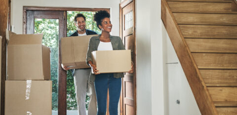 Couple carrying moving boxes into a home, stepping through the front door with more boxes nearby