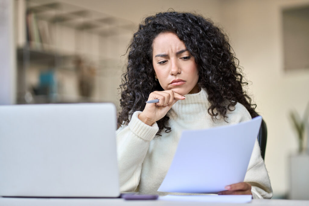 Person reviewing a document with a concerned expression while sitting at a desk with a laptop
