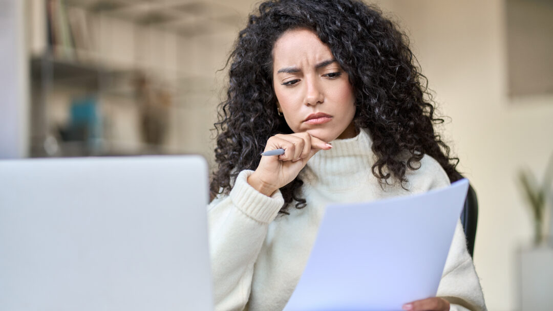 Person reviewing a document with a concerned expression while sitting at a desk with a laptop