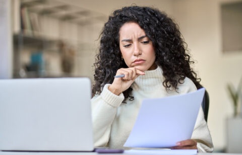 Person reviewing a document with a concerned expression while sitting at a desk with a laptop