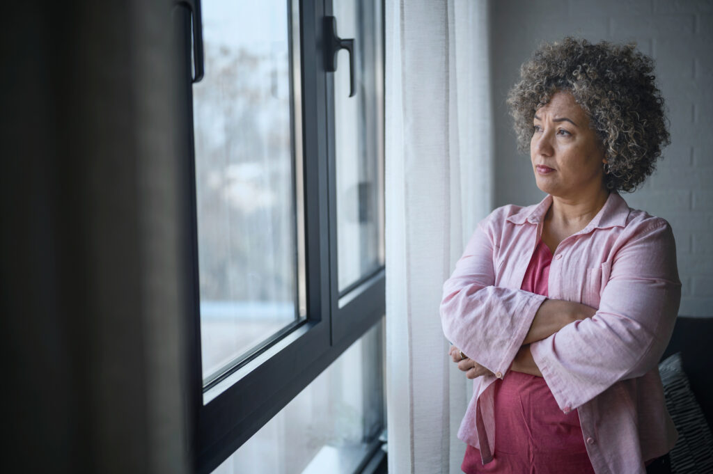 Woman standing by a window with her arms crossed, looking outside thoughtfully.