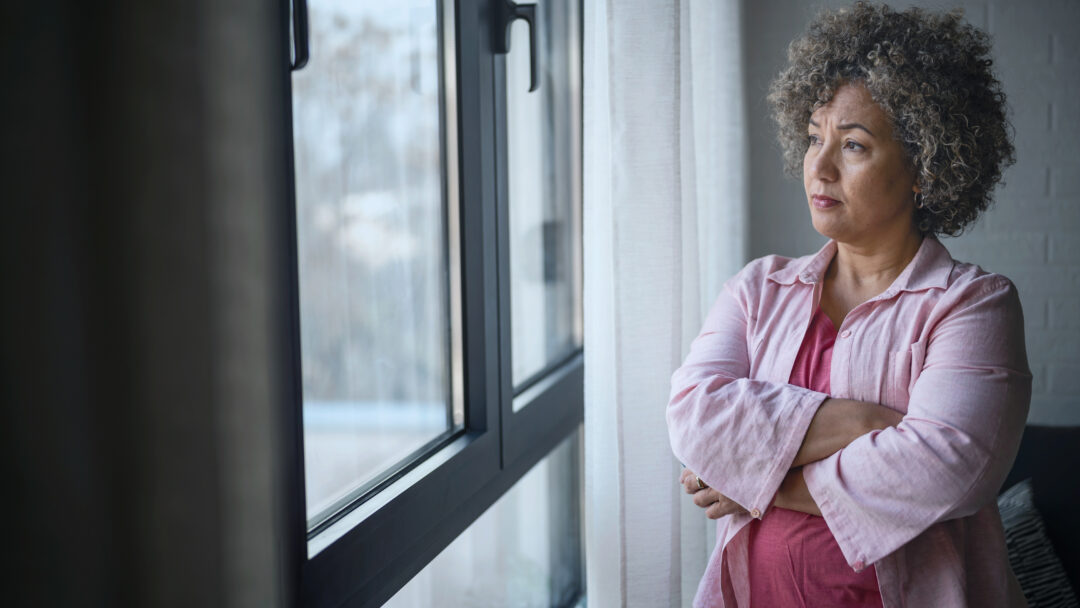 Woman standing by a window with her arms crossed, looking outside thoughtfully.