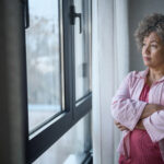 Woman standing by a window with her arms crossed, looking outside thoughtfully.