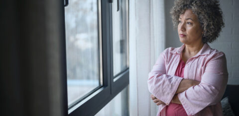 Woman standing by a window with her arms crossed, looking outside thoughtfully.
