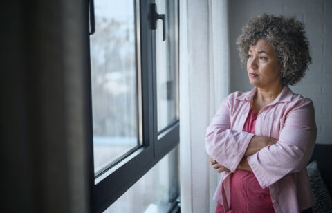 Woman standing by a window with her arms crossed, looking outside thoughtfully.