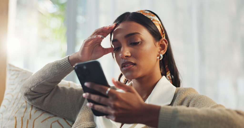 Person sitting on a couch looking at a smartphone, holding their head with a concerned expression