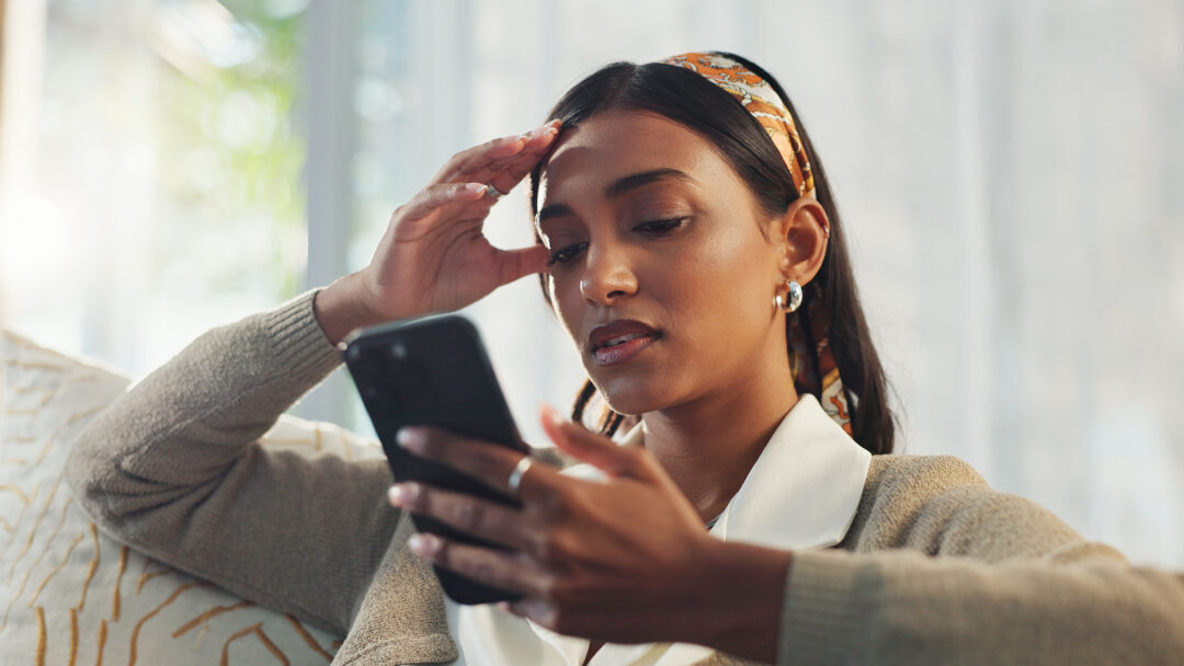 Person sitting on a couch looking at a smartphone, holding their head with a concerned expression
