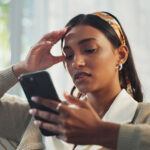 Person sitting on a couch looking at a smartphone, holding their head with a concerned expression
