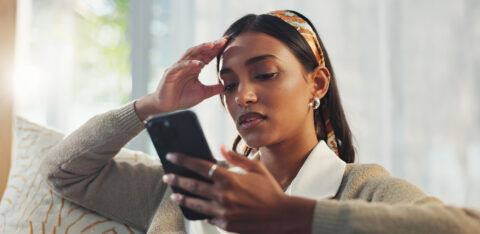 Person sitting on a couch looking at a smartphone, holding their head with a concerned expression