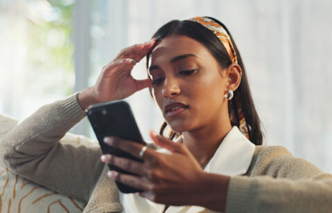 Person sitting on a couch looking at a smartphone, holding their head with a concerned expression