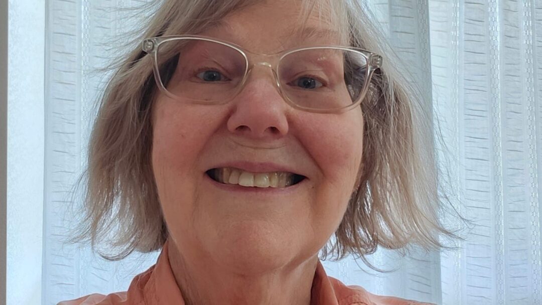 Close-up of a smiling older woman wearing glasses and a button-up shirt indoors