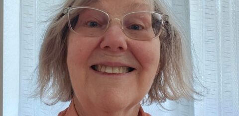 Close-up of a smiling older woman wearing glasses and a button-up shirt indoors