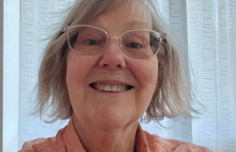 Close-up of a smiling older woman wearing glasses and a button-up shirt indoors