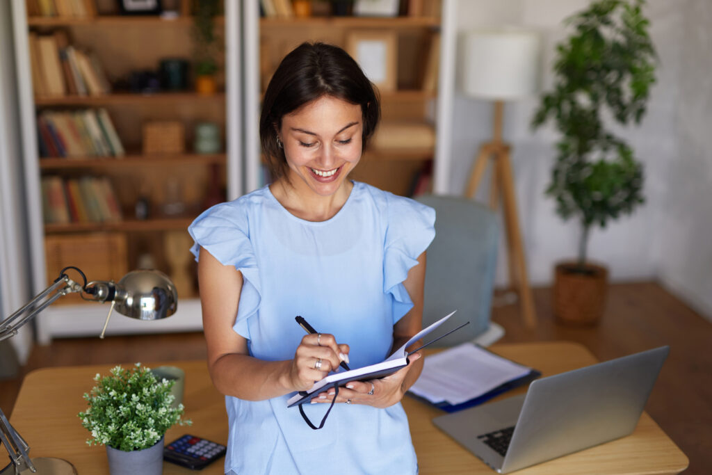 Smiling woman writing in a notebook at her home office desk with a laptop, calculator, and desk lamp nearby.