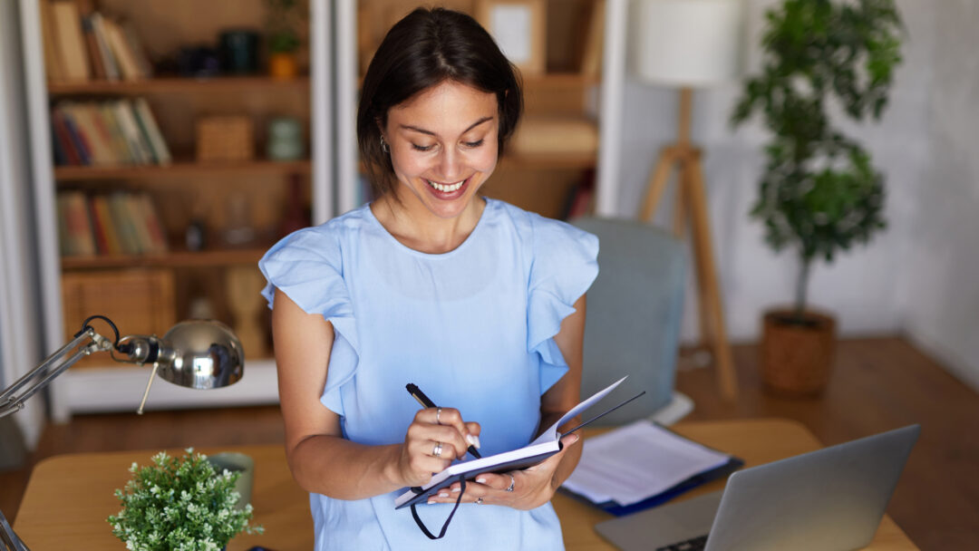 Smiling woman writing in a notebook at her home office desk with a laptop, calculator, and desk lamp nearby.