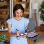 Smiling woman writing in a notebook at her home office desk with a laptop, calculator, and desk lamp nearby.