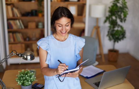 Smiling woman writing in a notebook at her home office desk with a laptop, calculator, and desk lamp nearby.