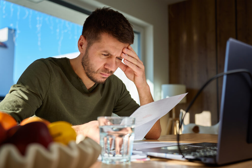 Man reviewing a bill at a table with a laptop, holding his forehead as he concentrates on his finances.