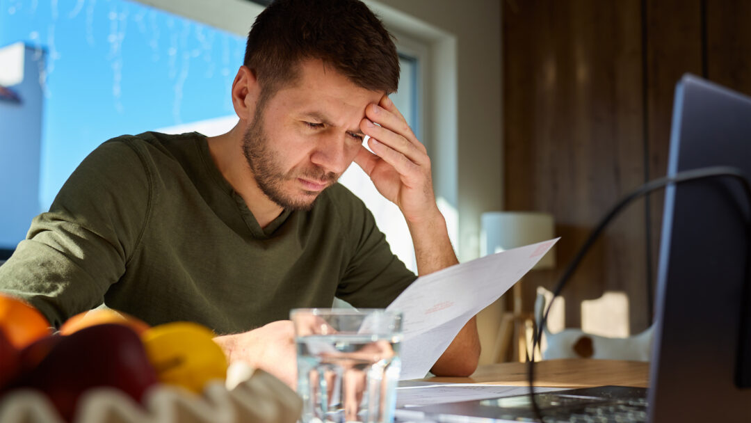 Man reviewing a bill at a table with a laptop, holding his forehead as he concentrates on his finances.
