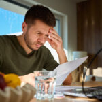 Man reviewing a bill at a table with a laptop, holding his forehead as he concentrates on his finances.