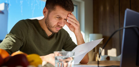 Man reviewing a bill at a table with a laptop, holding his forehead as he concentrates on his finances.