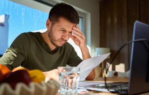 Man reviewing a bill at a table with a laptop, holding his forehead as he concentrates on his finances.