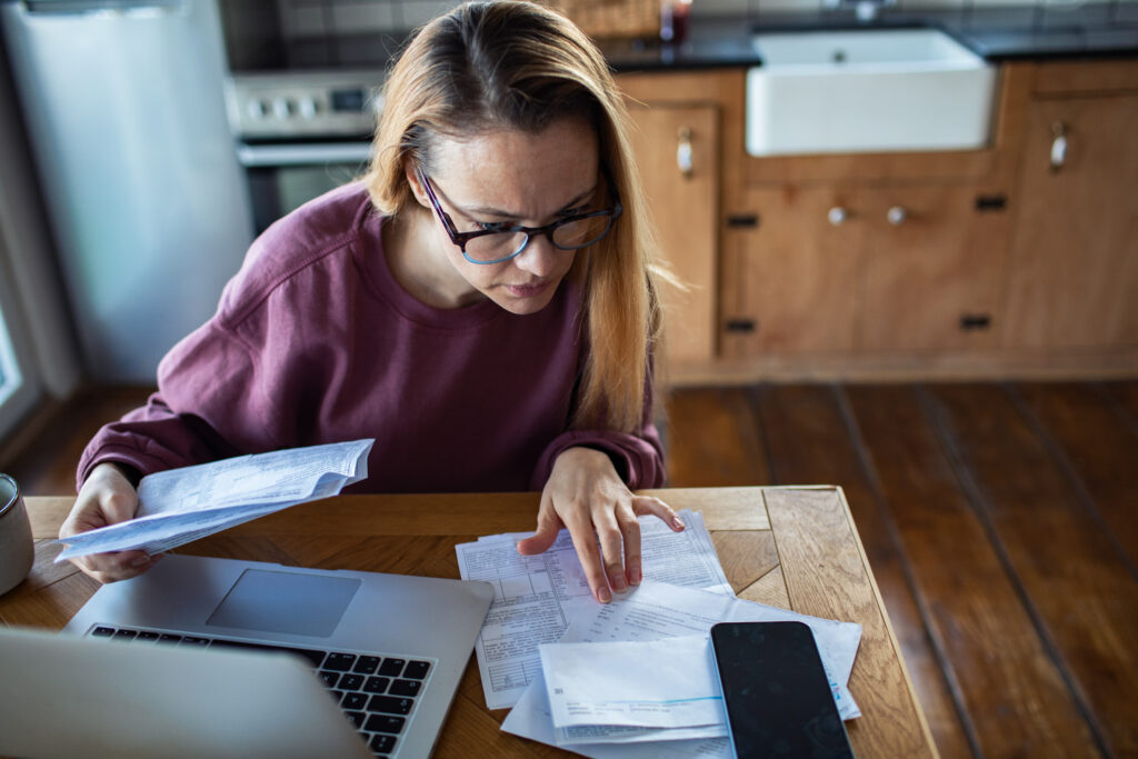 Woman reviewing bills and financial documents at a kitchen table with a laptop and smartphone, concentrating on her finances.