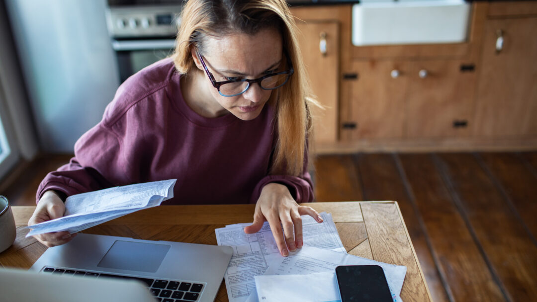 Woman reviewing bills and financial documents at a kitchen table with a laptop and smartphone, concentrating on her finances.