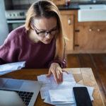 Woman reviewing bills and financial documents at a kitchen table with a laptop and smartphone, concentrating on her finances.