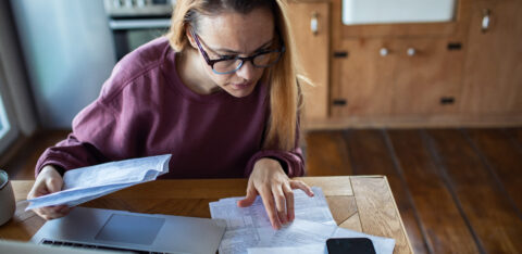 Woman reviewing bills and financial documents at a kitchen table with a laptop and smartphone, concentrating on her finances.