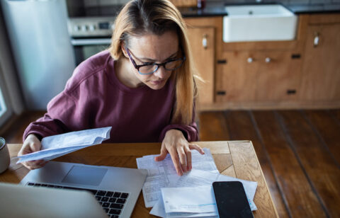 Woman reviewing bills and financial documents at a kitchen table with a laptop and smartphone, concentrating on her finances.
