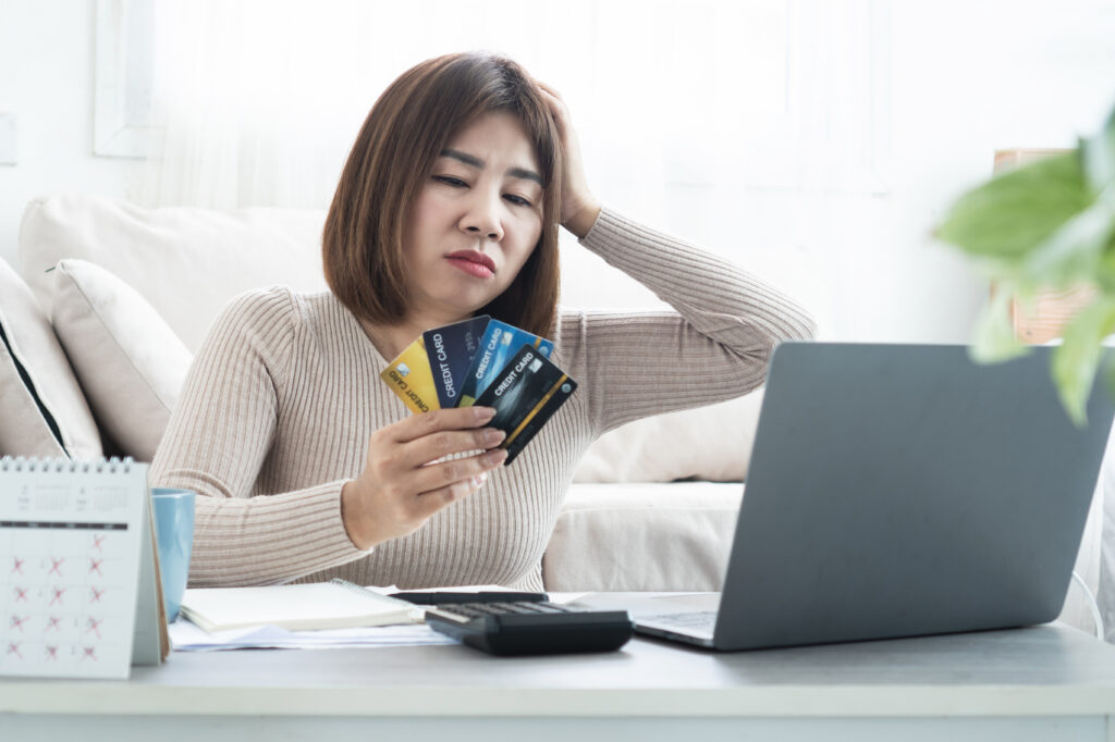 Concerned woman holding several credit cards while looking at a laptop, with a calculator, notebook, and calendar on the table.