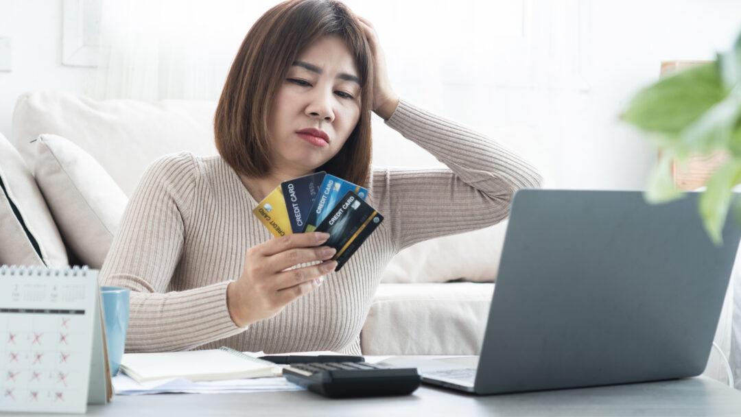 Concerned woman holding several credit cards while looking at a laptop, with a calculator, notebook, and calendar on the table.