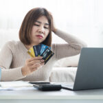 Concerned woman holding several credit cards while looking at a laptop, with a calculator, notebook, and calendar on the table.